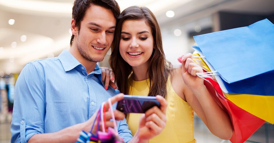 Pareja joven revisando un celular mientras sostiene bolsas de compras dentro de un centro comercial.